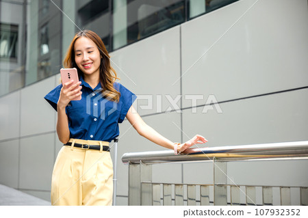 A young woman wearing blue shirt texting on her phone while strolling in the city. Girl in a good mood, busy with her smartphone app. 107932352