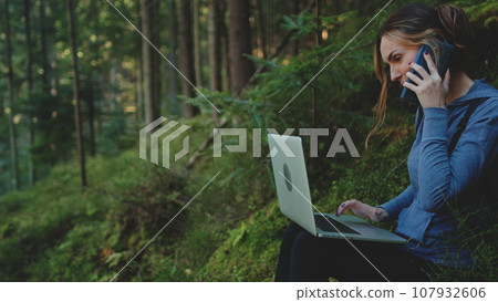 A woman working outdoors. Girl typing on laptop and talking by phone sitting in a green forest landscape. Digital nomad working on a laptop, remote working specialist working in nature with fresh air A woman working outdoors. Girl typing on laptop and talking by phone sitting in a green forest landscape. Digital nomad working on a laptop, remote working specialist working in nature with fresh air 107932606