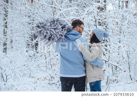 a loving couple in the winter forest is hugging. a man holds a Christmas tree on his shoulder 107933610