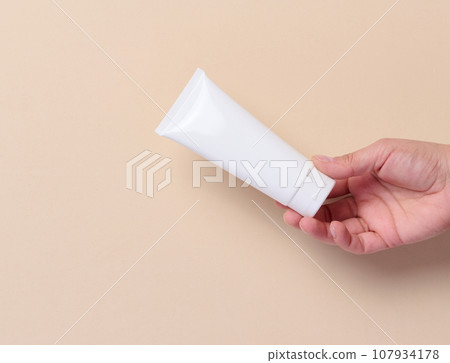 A woman's hand holds a white plastic tube for cream, toothpaste on a beige background 107934178