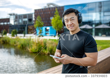 Portrait of a smiling young man, Asian athlete and runner standing on the street near the city wearing headphones and using the phone. 107934803