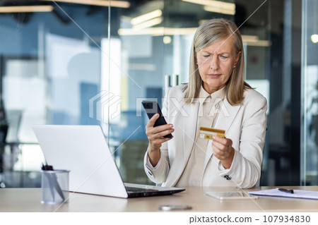 Problems with the financial account. Worried senior businesswoman sitting at desk in office, holding phone and looking confusedly at credit card. 107934830