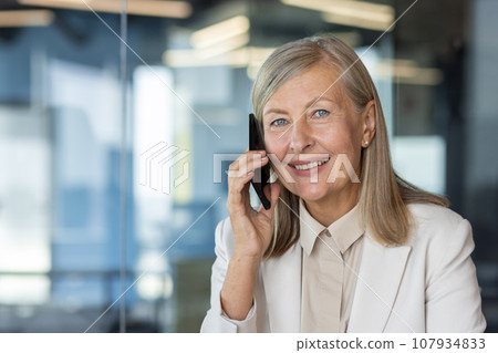 Close-up portrait of a smiling senior business woman in a suit sitting at a desk in the office and talking on a mobile phone. Close-up portrait of a smiling senior business woman in a suit sitting at a desk in the office and talking on a mobile phone. 107934833