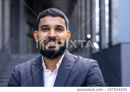 Close-up photo of a fashionable Muslim man, in a business suit standing outside on the street near a business center, and smiling at the camera. 107934838