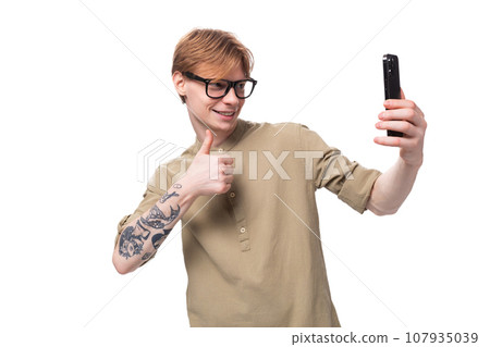 a young red-haired guy in glasses and a brown shirt takes a selfie on the phone 107935039