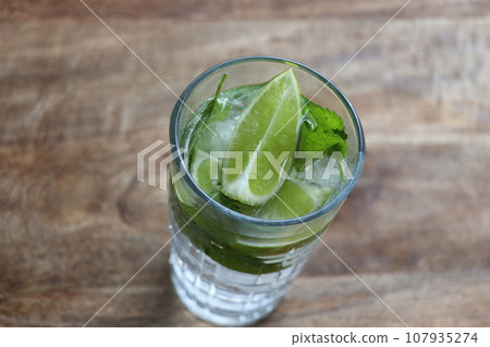 Glass of Mojito summer alcoholic cocktail with ice cubes mint and lime on wooden background Glass of Mojito summer alcoholic cocktail with ice cubes mint and lime on wooden background 107935274