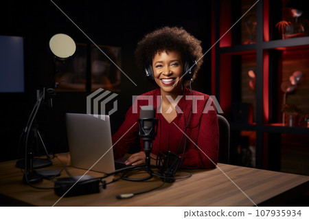 Young beautiful African American woman wearing headphones in a recording studio. Smiling black lady recording news, podcast or blog using microphone, laptop and professional studio equipment. Young beautiful African American woman wearing headphones in a recording studio. Smiling black lady recording news, podcast or blog using microphone, laptop and professional studio equipment. 107935934