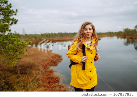Smiling female traveler in yellow coat walks along wooden path among wild nature. Travel, vacation. Smiling female traveler in yellow coat walks along wooden path among wild nature. Travel, vacation. 107937933