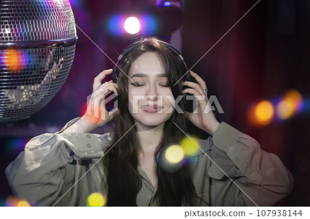 A beautiful girl in headphones enjoys music against the background of light rays from a disco ball. Disco, party. 107938144