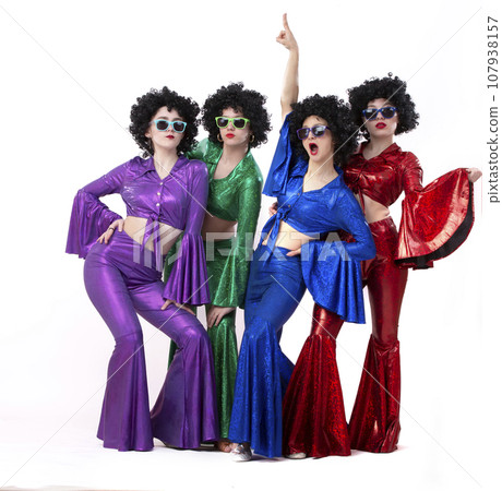 A group of girls in colorful flared suits and afro wigs pose against a white background. Disco style from the eighties or seventies. 107938157