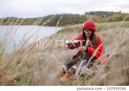 Woman holds thermos and drinks tea in autumn forest. Rest, relaxation, travel, lifestyle concept. 107938586