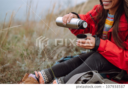 Woman holds thermos and drinks tea in autumn forest. Rest, relaxation, travel, lifestyle concept. Woman holds thermos and drinks tea in autumn forest. Rest, relaxation, travel, lifestyle concept. 107938587