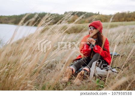 Woman holds thermos and drinks tea in autumn forest. Rest, relaxation, travel, lifestyle concept. Woman holds thermos and drinks tea in autumn forest. Rest, relaxation, travel, lifestyle concept. 107938588