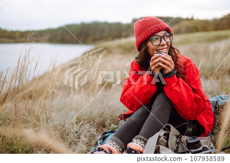 Woman holds thermos and drinks tea in autumn forest. Rest, relaxation, travel, lifestyle concept. Woman holds thermos and drinks tea in autumn forest. Rest, relaxation, travel, lifestyle concept. 107938589