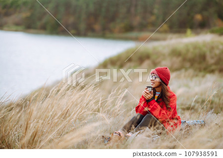 Woman holds thermos and drinks tea in autumn forest. Rest, relaxation, travel, lifestyle concept. Woman holds thermos and drinks tea in autumn forest. Rest, relaxation, travel, lifestyle concept. 107938591