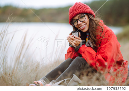 Woman holds thermos and drinks tea in autumn forest. Rest, relaxation, travel, lifestyle concept. Woman holds thermos and drinks tea in autumn forest. Rest, relaxation, travel, lifestyle concept. 107938592