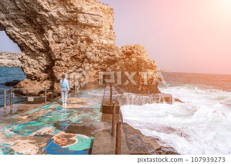 woman sea travel. A woman in a blue jacket stands on a rock above a cliff above the sea, looking at the stormy ocean. Girl traveler rests, thinks, dreams, enjoys nature 107939273