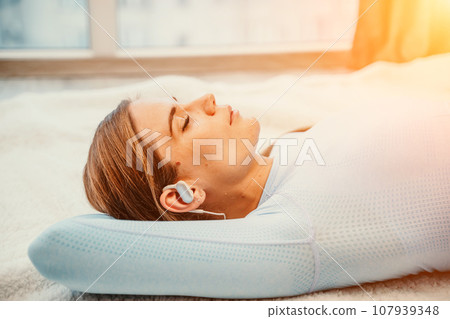 Side view portrait of relaxed woman listening to music with headphones lying on carpet at home. She is dressed in a blue tracksuit. Side view portrait of relaxed woman listening to music with headphones lying on carpet at home. She is dressed in a blue tracksuit. 107939348