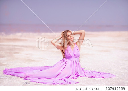 Pink lake woman. A woman in a pink dress sits on the salty shore of a pink lake and poses for a souvenir photo, creating lasting memories. 107939369