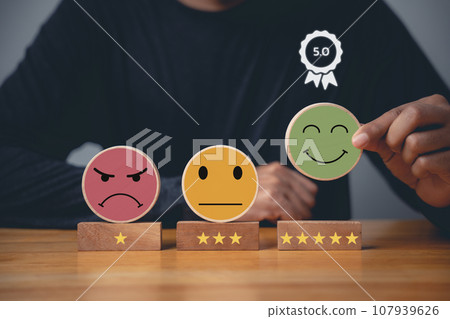 Hand choosing a smiley face on a wooden block circle to rate the best business services and customer experience. Satisfaction survey and 5-star rating concept. 107939626