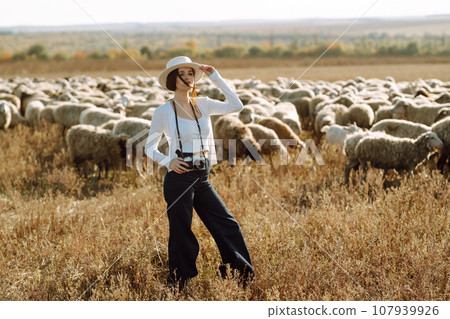 Young woman with a camera in front of a herd of sheep. Nature, fashion, vacation and lifestyle. 107939926