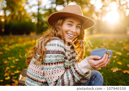 Curly woman with thermos in clearing among fallen leaves in autumn park, enjoying hot drink. Curly woman with thermos in clearing among fallen leaves in autumn park, enjoying hot drink. 107940108