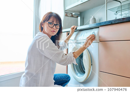 Middle aged woman near washing machine in kitchen at home Middle aged woman near washing machine in kitchen at home 107940476