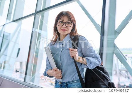 Woman waiting for airplane flight in airport terminal, holding laptop Woman waiting for airplane flight in airport terminal, holding laptop 107940487