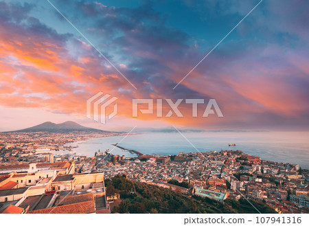 Naples, Italy. Top View Skyline Cityscape City In Evening Sunset. Tyrrhenian Sea And Landscape With Volcano Mount Vesuvius. Amazing Sunset Sky With Saturated Colorful Clouds. Sun Shining With Sun Rays 107941316
