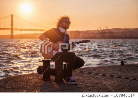 Hipster street musician in black playing electric guitar in the street on sunset on embankment with 25th of April bridge in background. Lisbon, Portugal Hipster street musician in black playing electric guitar in the street on sunset on embankment with 25th of April bridge in background. Lisbon, Portugal 107945114