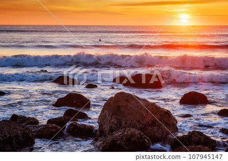 Atlantic ocean sunset with waves and rocks and surfers silhouettes in water at Costa da Caparica, Portugal 107945147