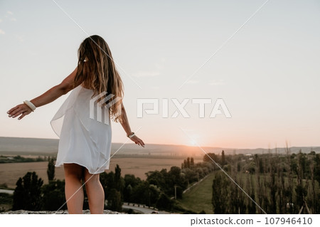 Happy woman standing with her back on the sunset in nature in summer with open hands. Romantic beautiful bride in white boho dress posing with mountains on sunset Happy woman standing with her back on the sunset in nature in summer with open hands. Romantic beautiful bride in white boho dress posing with mountains on sunset 107946410