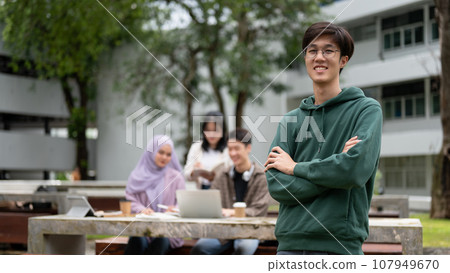 A smart male college student stands with his arms crossed while his friends are working on a project A smart male college student stands with his arms crossed while his friends are working on a project 107949670