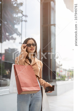 Gorgeous and confident Asian woman in trendy clothes wearing sunglasses, carrying her shopping bags Gorgeous and confident Asian woman in trendy clothes wearing sunglasses, carrying her shopping bags 107949781