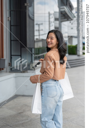 An attractive Asian woman is walking outside of a shopping mall with her shopping bags. 107949787