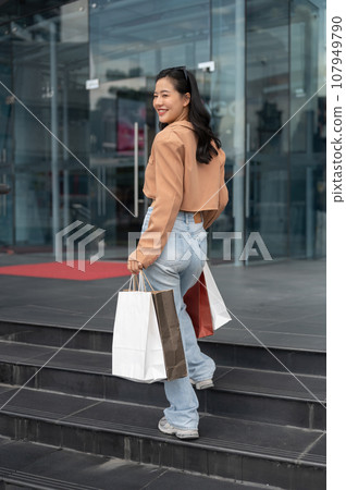 A joyful Asian woman is getting into a shopping mall, walking with her shopping bags. A joyful Asian woman is getting into a shopping mall, walking with her shopping bags. 107949790