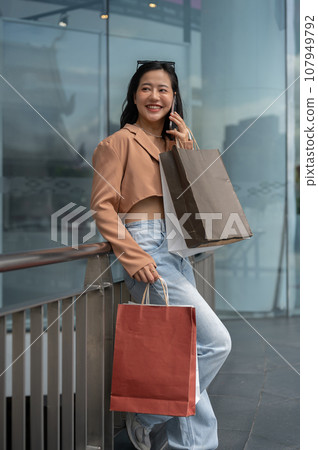 A trendy Asian woman is talking on the phone with her friend during her shopping day at the mall. 107949792