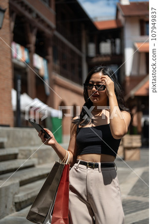 Gorgeous Asian woman enjoys shopping in the city, walking while holding her phone and shopping bags Gorgeous Asian woman enjoys shopping in the city, walking while holding her phone and shopping bags 107949837