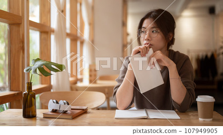 A pensive, thoughtful young Asian woman sits at a table in a beautiful, cosy coffee shop. A pensive, thoughtful young Asian woman sits at a table in a beautiful, cosy coffee shop. 107949905