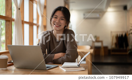A charming Asian woman sitting at a table with her laptop and books in a minimal coffee shop. 107949917