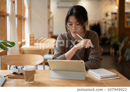 A focused Asian woman is working on her assignment, using a digital tablet in a cosy cafe. 107949922