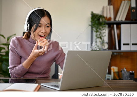 Beautiful young Asian girl eating a donut while studying at her study table Beautiful young Asian girl eating a donut while studying at her study table 107950130