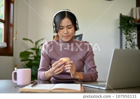 Beautiful young Asian girl eating a donut while studying online at her study table 107950131