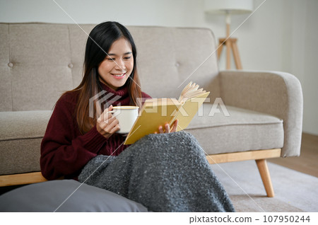 A happy Asian woman is sipping hot chocolate and reading a book in her living room A happy Asian woman is sipping hot chocolate and reading a book in her living room 107950244