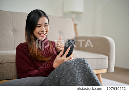 A cheerful Asian woman in a cozy sweater enjoys talking on a video call while relaxing at home 107950252