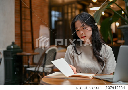 A beautiful Asian woman focuses on reading a book while working remotely at a coffee shop. 107950334