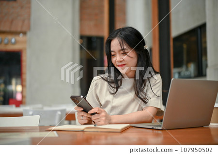 A pretty Asian woman is texting with her friends while working remotely at a coffee shop. A pretty Asian woman is texting with her friends while working remotely at a coffee shop. 107950502