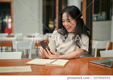 A pretty Asian woman is texting with her friends while working remotely at a coffee shop. A pretty Asian woman is texting with her friends while working remotely at a coffee shop. 107950503