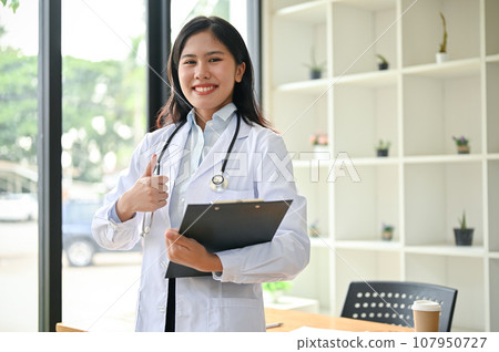A beautiful Asian female doctor in a white gown stands and shows her thumb up to the camera. A beautiful Asian female doctor in a white gown stands and shows her thumb up to the camera. 107950727