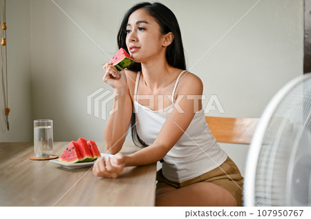 A beautiful Asian woman eats a slide of watermelon and sits in front of an electric fan 107950767